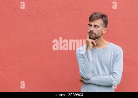 Young bearded man in thoughtful pose with fingers on chin on red wall background with copy space. Minded man trying to find way to solve problem. Doub Stock Photo