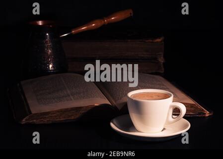 Cup of hot coffee, copper cezve and open Torah page on black wood table in the dark. Closeup Stock Photo