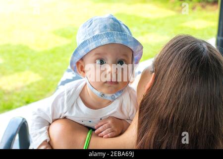 New mother and child on deckchair at backyard with green grass garden view and holding her little baby, baby boy resting on mother’s shoulder and look Stock Photo