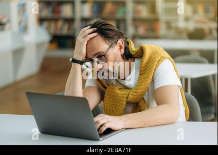 Frustrated male manager is sitting at the work desk, sed about failed project. Middle aged businessman using laptop, having a terrible headache, feeling tired, overwork concept Stock Photo
