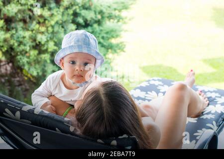 Beautiful mother holding curious baby boy in hat while lays on flower pattern deck chair, enjoying maternity outside Stock Photo