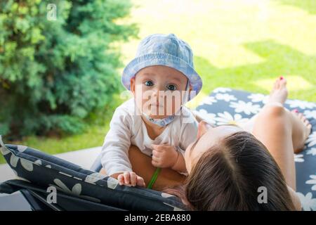 Young mother with little baby in sun cap laying on flower pattern deck chair and playing outdoor together, green natural backyard view on background Stock Photo