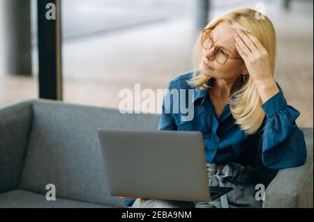 Senior adult business woman is sitting on the couch, using laptop. Exhausted female employee is having a terrible headache, feeling bad after tense brainstorm, overwork concept Stock Photo