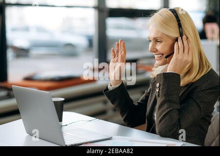 Middle aged female employee sit at the work desk, having a video conference with colleagues, waves hand and greeting. Friendly charming woman wearing headset, works as employee of support line, smiles Stock Photo