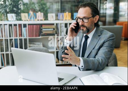 Shocked businessman using laptop and smartphone, having a serious conversation, sees on the laptop screen unexpected news. Senior adult male employee got surprised message or having problem Stock Photo