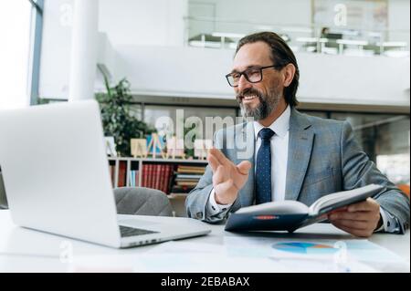 Smiling male employee wearing in formal suit, using laptop, makes notes at online conference.Mature successful businessman sits at the desk, working remotely, communicate with colleagues by video call Stock Photo