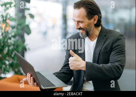 Successful businessman is sitting on the sofa, using laptop, making online conference, discussing new strategy for company. Joyful male manager approved a work project on video briefing Stock Photo