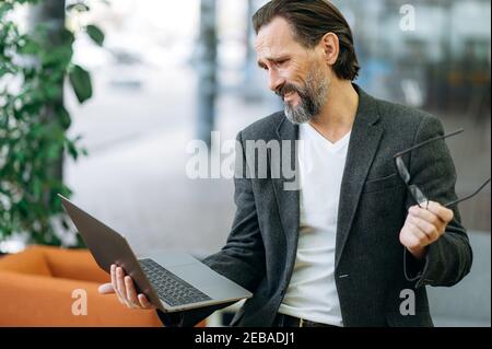 Confused male employee sit on the sofa in office, using laptop, having problems with internet connection. Senior adult businessman in formal suit upset with failed project, unsuccess concept Stock Photo