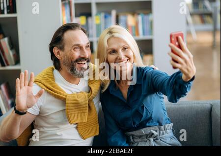 Happy senior business partners using smartphone, having a video call from colleagues. Successful coworkers sitting on the sofa in office while break, talking about work process, communication concept Stock Photo