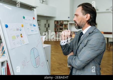 Middle aged businessman is working on important project, brainstorming. Pensive male employee stands in office, looks at the flipchart, thinking about ideas for startup, learning financial graphs Stock Photo