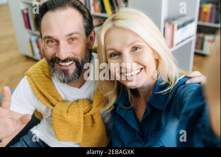 Middle aged joyful colleagues using smartphone, doing a selfie, taking a break from work. Friendly business people in stylish wear looking directly at the camera, smiling, partnership concept Stock Photo