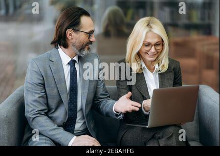 Confident middle aged business partners sitting on the sofa, discussing project, smiling. Male manager and elegant female employee using laptop, searching ideas, browsing internet, teamwork concept Stock Photo