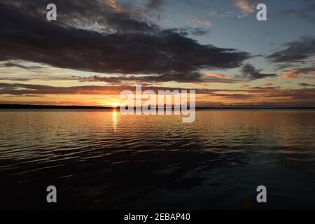 Sunbeam in the sky from under a dark cloud of the setting sun on the horizon above the water on sunset Stock Photo