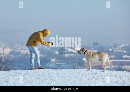 Pet owner taking photo of his labrador retriever via smart phone during frosty day. Young man with dog in winter against urban skyline. Prague, Czech Stock Photo