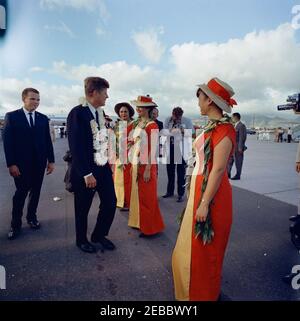 Trip to Western States: Honolulu, Hawaii, departure from Honolulu International Airport, 5:35PM. President John F. Kennedy (left, wearing a flower lei) stands on the tarmac before departing from Honolulu International Airport in Honolulu, Hawaii; several woman (wearing red and yellow dresses with matching hats) stand in a row at center. White House Secret Service agent, Ron Pontius, stands at far left. Stock Photo