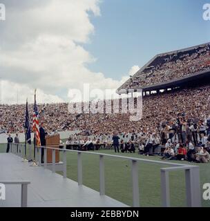 Inspection tour of NASA installations: Houston, Texas, motorcade, address at Rice University, 9:34AM. President John F. Kennedy (left, at lectern) delivers remarks at Rice University regarding the nationu0027s efforts in space exploration. White House Secret Service agent, Roy Kellerman, walks at far left. Members of the press sit in background; photographers gather at right; spectators observe from the stands. Rice University Stadium, Houston, Texas. Stock Photo