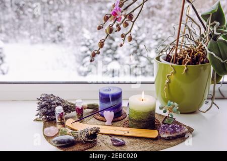 Small good feng shui altar in home on window sill on leaf shape table mat, snowy Nordic nature on background. Incense candle smoking, gemstones. Stock Photo