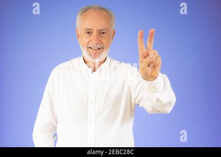 Senior grey-haired man wearing casual shirt standing over isolated violet background smiling with happy face looking at the camera doing victory sign Stock Photo