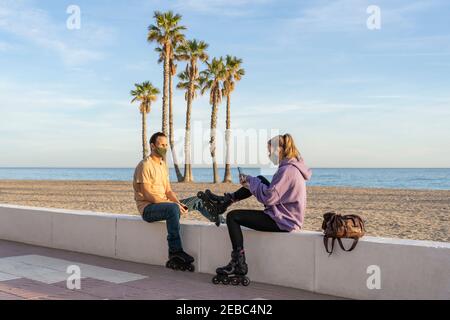 Skateboarding couple in medical protective mask sitting on promenade,using cell phone on seaside, palm trees and beach background.Healthy , fitness te Stock Photo