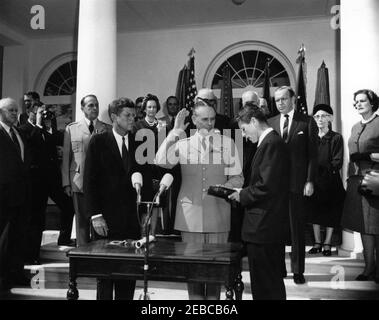 Swearing-in ceremony, Gen. Maxwell Taylor, Chairman, JCS, 12:00PM. President John F. Kennedy (center left) and others observe as General Maxwell D. Taylor (with right hand raised and left hand on Bible) is sworn in as Chairman of the Joint Chiefs of Staff (JCS); Attorney General, Robert F. Kennedy (center right, holding Bible), administers the oath of office. Looking on from the steps of the West Wing Colonnade, beginning at the very back (left to right): Naval Aide to the President, Captain Tazewell T. Shepard, Jr.; General Omar Bradley; unidentified motion picture photographer; outgoing Chai Stock Photo