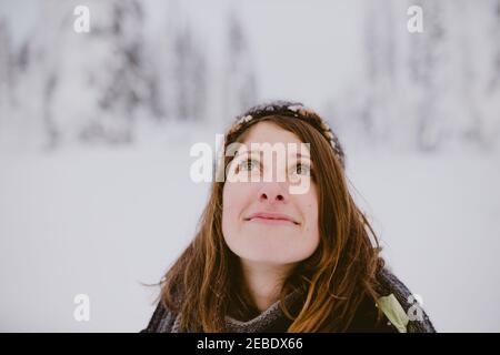 Young woman with brown hair and eyes looks up at snow covered trees Stock Photo