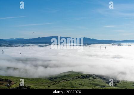 A view of rolling hills landscape in Andalusia with many wind turbines above the fog in the valleys and blue sky above Stock Photo