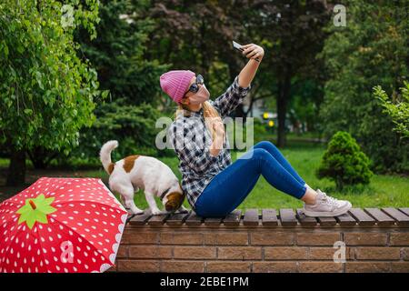 Portrait of a young woman making selfie photo on smartphone sitting on the bench with dog in the park Stock Photo