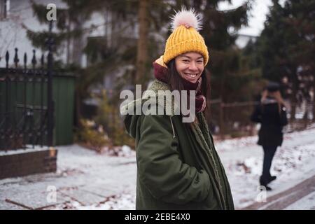 Happy Asian woman with yellow fluffy beanie smiles in snowy street Stock Photo
