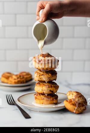 Hand pouring icing on fresh cinnamon buns stacked high on a plate. Stock Photo