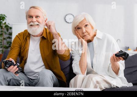 KYIV, UKRAINE - DECEMBER 17, 2020: Excited senior man holding joystick near sad wife at home Stock Photo