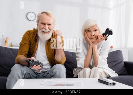 KYIV, UKRAINE - DECEMBER 17, 2020: Senior man with joystick showing yes gesture near sad wife Stock Photo