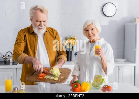 Senior woman holding glass of orange juice near husband pouring vegetables in bowl in kitchen Stock Photo