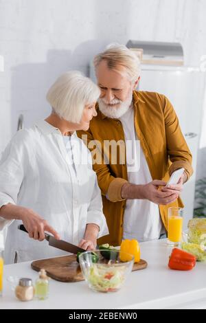 Senior man holding smartphone near smiling wife cooking on blurred foreground in kitchen Stock Photo