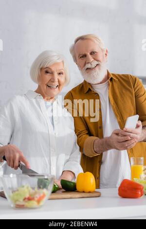 Smiling senior couple with smartphone looking at camera while cooking on blurred foreground Stock Photo