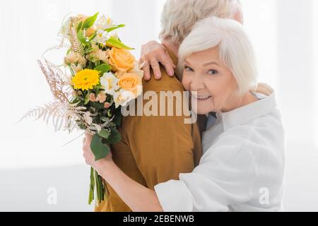 https://l450v.alamy.com/450v/2ebenwb/smiling-senior-woman-embracing-husband-and-holding-bouquet-at-home-2ebenwb.jpg