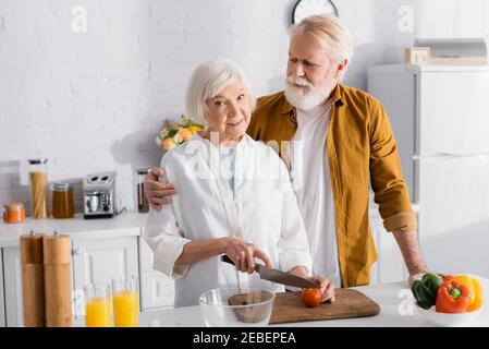 Elderly man embracing wife cutting tomato while cooking in kitchen Stock Photo