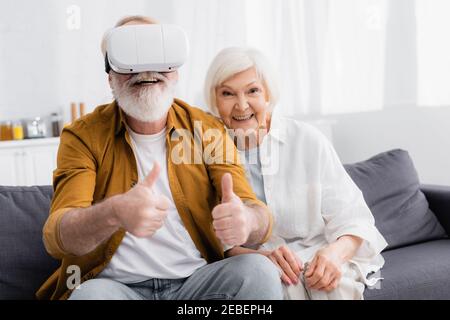 Senior man in virtual reality headset showing like near smiling wife at home Stock Photo
