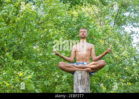 Young man sitting on tree trunk and meditating with closing eyes and his hands turning up to sky on foliage background. Guy sitting in lotus pose on t Stock Photo