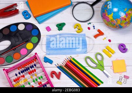 Back to school flat lay, top view concept. School supplies, pencils, paints, globe, scissors, colored books, alphabet and medical mask on the white ta Stock Photo