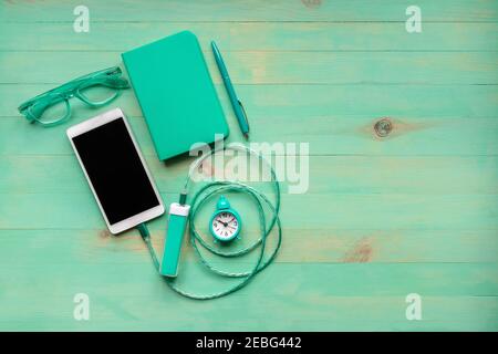 Charging smartphone with portable external battery, alarm clock, paper notebook, pen and eyeglasses on wooden desk, all in turquoise color. Phone char Stock Photo