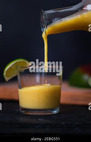 A vibrant yellow orange fruit smoothie or juice being poured into a clear glass with lime segment on the rim. Fruit in bokeh background against dark s Stock Photo