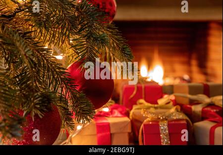 Xmas home celebration concept. Christmas tree branch with red ornament balls, gift boxes with ribbons, bows and blur burning fireplace background. War Stock Photo