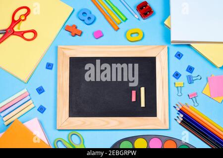 Back to school flat lay composition with blackboard. School supplies, pencils, paints, scissors, magnifying glass and colored books, on blue background Stock Photo