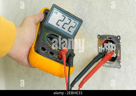 Male electrician checks the voltage of an outlet on a white wall with a multimeter in the house close up Stock Photo