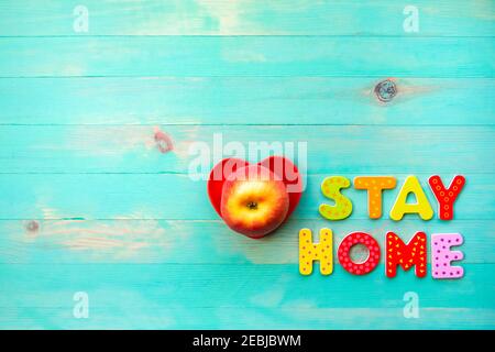 Words STAY HOME and apple in red heart shape plate on turquoise wooden background. Text STAY HOME of wooden letters and fruit in bowl top view. Corona Stock Photo