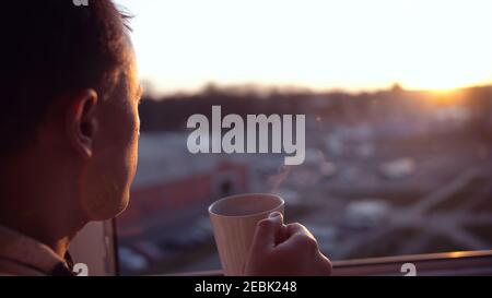 A man stands at the window and looks at the sunset, drinks tea from a mug, close-up Stock Photo