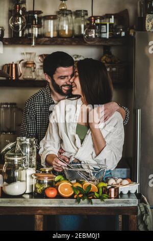 Sweet young family couple cookin together in the kitchen. Young caucasian man and brunette woman with faces stained in flour hugging, laughing and making dough for baking citrus cake. Valentines Day, home hobbies concept Stock Photo