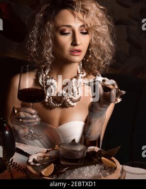 Portrait of beautiful blonde curly hair woman in seafood restaurant eating oysters holding one in hand and drink wine Stock Photo