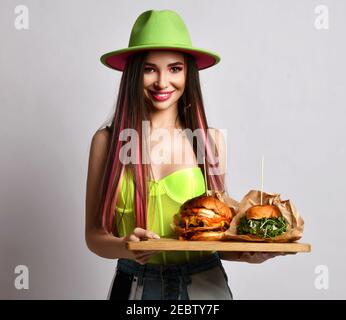 Young woman in jeans, corset and wide-brimmed hat stands holding two craft big burger sandwiches on wooden tray
