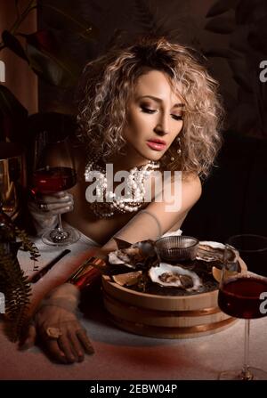 Portrait of beautiful blonde curly hair woman in seafood restaurant eating oysters and drink wine on dark Stock Photo
